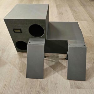 May include: A grey cat house with a brick pattern, featuring two circular openings and a rectangular window. The structure includes two rectangular support legs. The cat house is designed to provide a cosy and stylish space for feline companions.