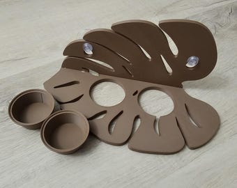 May include: A brown, leaf-shaped organizer with two round cup holders and two suction cups. The organizer is designed to hold small items. The leaf design has cut-out circles and curved edges. The organizer is made of a solid material.