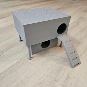 以下が含まれることがあります： 質感のある素材で作られた、グレーの2階建ての猫用ハウスとスロープ。ハウスには丸い開口部が2つあり、スロープには5つのステップがあります。