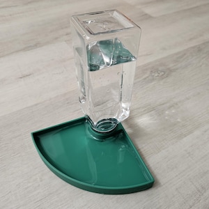 May include: A green plastic pet water dispenser with a clear glass bottle filled with water. The bottle is attached to a green plastic base with a rounded edge.