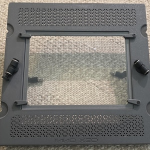 May include: A gray rectangular frame with a clear glass center. The frame has a perforated top and bottom section, with black fittings on the sides. The text "INNO 3D" is visible on the bottom right corner. The frame is designed for a computer component.