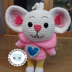 Peut inclure: Poupée souris au crochet blanc faite à la main, avec de grandes oreilles avec des accents de cœur roses. La souris porte un pull rose avec un cœur bleu appliqué et un pantalon rayé jaune et blanc. La poupée a des yeux noirs et un nez rose.