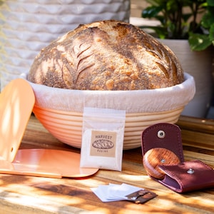 May include: A set of bread baking tools including a proving basket, a dough scraper, a lame, a small bag of flour, and a leather case with a wooden scoring tool. The tools are arranged on a wooden surface.
