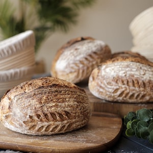 Three freshly baked sourdough loaves with deep golden crusts and intricate scoring, resting on wooden boards. Stacks of oval banneton proofing baskets with linen liners are visible in the background, highlighting their role in shaping artisan bread.