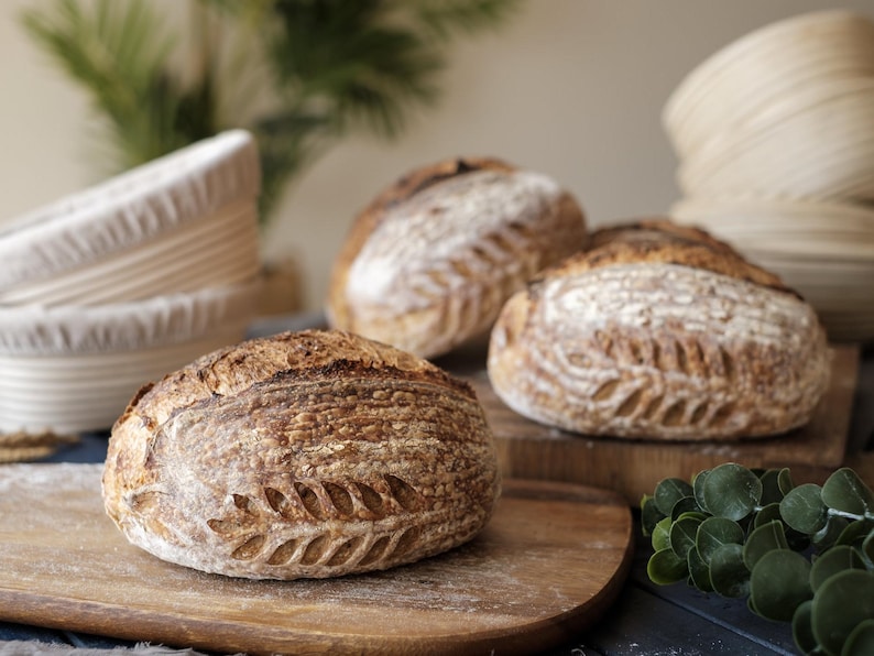 Three freshly baked sourdough loaves with deep golden crusts and intricate scoring, resting on wooden boards. Stacks of oval banneton proofing baskets with linen liners are visible in the background, highlighting their role in shaping artisan bread.