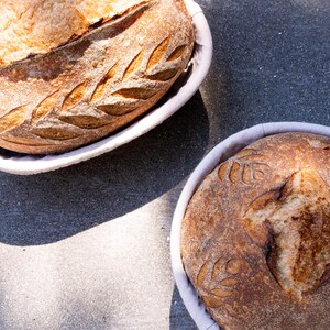 Two artisan sourdough loaves with decorative scoring, resting in oval and round banneton proofing baskets lined with linen. The crusts are golden and crisp, showcasing the texture and patterns created during the proofing process.