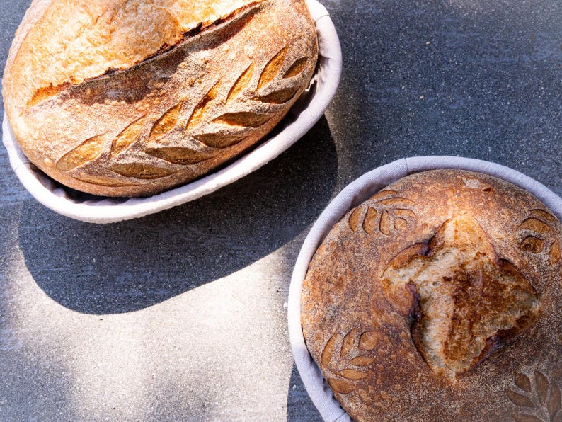 Two artisan sourdough loaves with decorative scoring, resting in oval and round banneton proofing baskets lined with linen. The crusts are golden and crisp, showcasing the texture and patterns created during the proofing process.