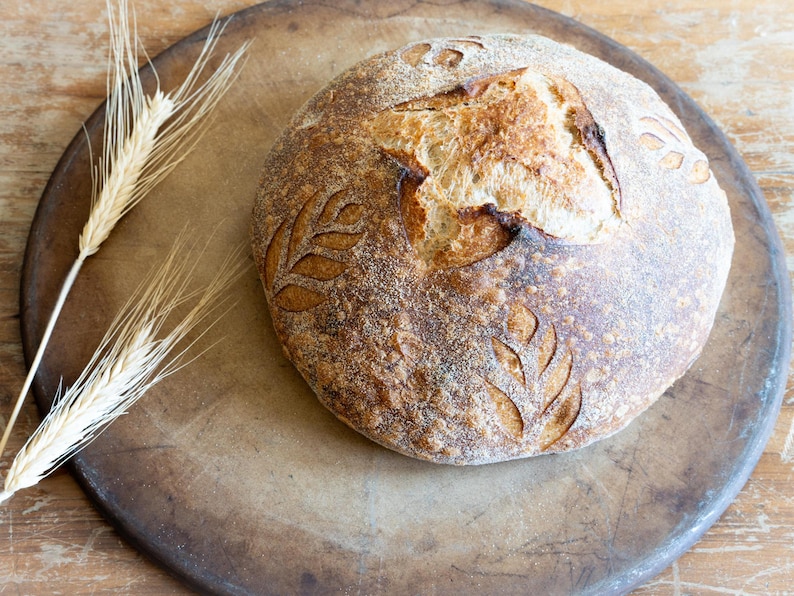 A round artisan sourdough loaf with decorative leaf scoring, resting on a well-used baking stone. The golden crust has a crisp texture, and wheat stalks are placed beside it on a rustic wooden surface.