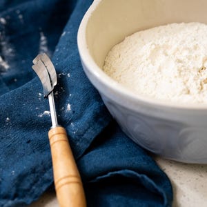 May include: A blue cloth with white flour scattered on it, a wooden handled dough cutter, and a white ceramic bowl filled with white flour.