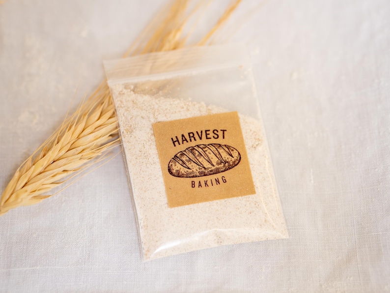 A packet of dehydrated sourdough starter from Harvest Baking, sealed in a clear plastic bag with a kraft paper label featuring a hand-drawn loaf illustration. Placed on a linen cloth with wheat stalks, showcasing the natural fermentation culture used for artisan bread baking.