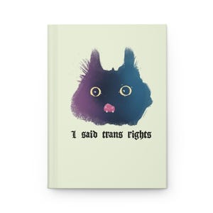 May include: A light green journal with the text "I said trans rights" in black gothic font. The cover features a stylized black cat face with large yellow eyes and a pink tongue, with purple and blue accents.