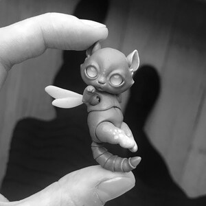 Woman holding a tiny 5 cm (2 inches) ball jointed cat between her thumb and first finger. Cat has big reflective eyes and a white scarf. Black and white photo.