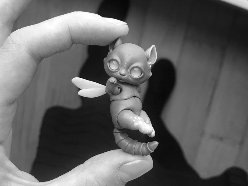Woman holding a tiny 5 cm (2 inches) ball jointed cat between her thumb and first finger. Cat has big reflective eyes and a white scarf. Black and white photo.