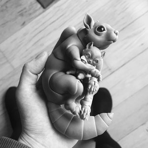 B&W photo of woman holding a ball jointed 3D printed squirrel with big black eyes and trusting face. It holds a cat doll with big reflective gray eyes and a white scarf.