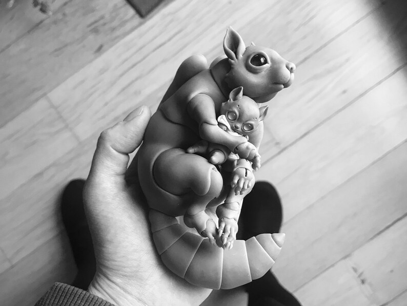 B&W photo of woman holding a ball jointed 3D printed squirrel with big black eyes and trusting face. It holds a cat doll with big reflective gray eyes and a white scarf.