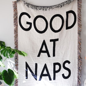 Può includere: Una coperta in tessuto bianco tessuta con frange nere e il testo "GOOD AT NAPS" in lettere nere.