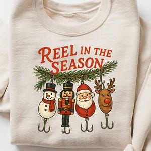 May include: Cream-colored sweatshirt with the text "REEL IN THE SEASON" in red. The design features four Christmas-themed fishing lures: a snowman, a nutcracker, Santa Claus, and a reindeer, all hanging from a green pine branch.