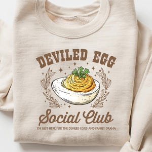 May include: Cream-coloured sweatshirt featuring the words "Deviled Egg Social Club" and a deviled egg graphic. The text "I'm just here for the deviled eggs and family drama" is below the club name.
