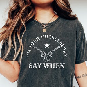 May include: Dark grey t-shirt featuring the text "I'm your Huckleberry, Say When" in white, with a star and crossed pistols design. The shirt is worn with blue jeans and white trainers. The theme is Western.