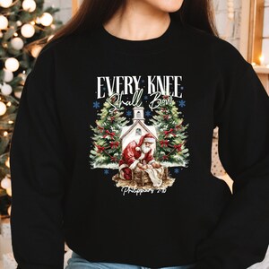 May include: Black sweatshirt with a Christmas-themed graphic. The graphic features Santa Claus, a church, and text that reads "EVERY KNEE SHALL BOW" above the image and "Philippians 2:10" below. A Christmas tree with lights is in the background.