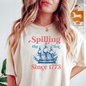 May include: Cream-colored t-shirt with the text "Spilling the Tea Since 1773" in pink and blue. The design features a blue illustration of a tall ship. The shirt is a casual, comfortable style.