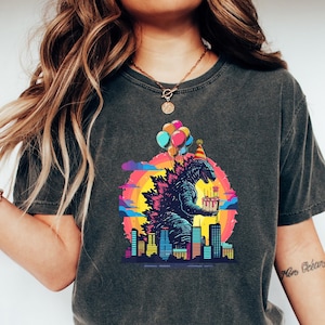 May include: A dark gray t-shirt with a graphic print of a Godzilla-like monster holding a birthday cake and wearing a party hat. The monster is surrounded by a cityscape and balloons.