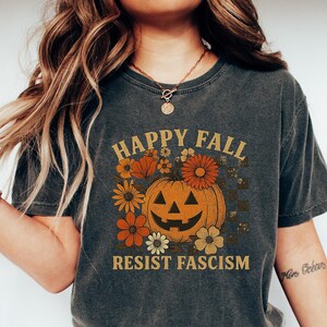 May include: Dark gray t-shirt with a vintage-style design. The shirt features the text "HAPPY FALL RESIST FASCISM" with a smiling jack-o'-lantern and floral accents in orange and cream colors. The shirt is paired with blue jeans and white sneakers.
