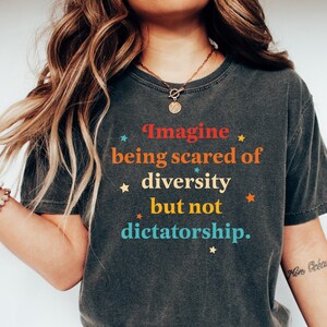 May include: A woman wearing a dark gray t-shirt with the text "Imagine being scared of diversity but not dictatorship." printed on it. The text is in a colorful font with stars around it. She is wearing blue jeans and white sneakers.