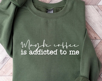May include: An olive green sweatshirt with the white text "Maybe coffee is addicted to me". The long-sleeved crew neck top is made of a soft material and is laid flat on a wooden surface.