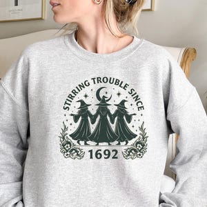 May include: A light gray sweatshirt with a graphic featuring three witches, a crescent moon, stars, and the text "Stirring Trouble Since 1692." The design is in dark green and white.