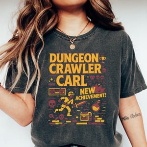 May include: A charcoal grey t-shirt with a graphic design featuring a pixelated character running with a sword, a treasure chest, and the text "Dungeon Crawler Carl New Achievement!"