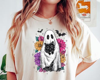 May include: Cream-colored t-shirt featuring a black and white illustration of a ghost holding a black cat. The design includes colorful flowers, bats, and stars. A casual, comfortable tee for Halloween or everyday wear.