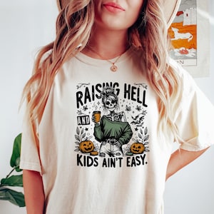 May include: Cream-colored t-shirt with a graphic featuring a skeleton holding a coffee cup, wearing a green sweater, and surrounded by pumpkins and bats. The text reads "RAISING HELL AND KIDS AIN'T EASY."
