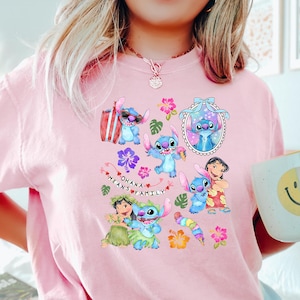 Peut inclure: Un t-shirt rose clair avec un motif color&eacute; de Stitch de Lilo & Stitch, entour&eacute; de fleurs et du texte "Ohana Means Family". Le motif comprend diverses images de Stitch. Une tasse blanche avec un smiley jaune est tenue dans la main.