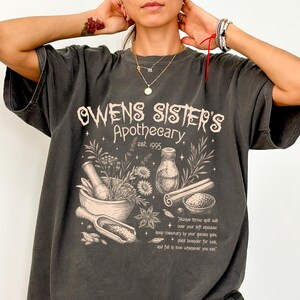 May include: A charcoal gray short-sleeve t-shirt featuring a cream-colored graphic print that reads Owens Sister's Apothecary est. 1995. The design includes illustrations of a mortar and pestle, a glass bottle, cinnamon sticks, and herbs.