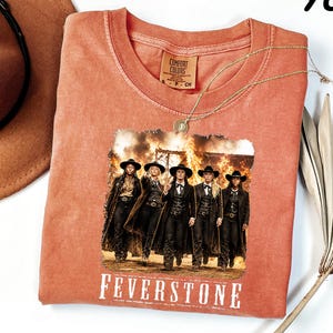 May include: A yam-colored t-shirt featuring a graphic print of five individuals in western attire walking in front of a large fire. The text "FEVERSTONE" is printed in white below the graphic. The shirt has a Comfort Colors brand label.