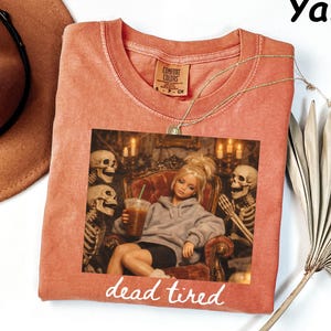 May include: A yam-colored t-shirt featuring a graphic print of a doll in a grey hoodie holding an iced coffee, flanked by two skeletons in a room with candles. The text "dead tired" is printed in white below the graphic, and the shirt has a tag.
