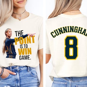 May include: A cream-colored t-shirt featuring a graphic of a basketball player in a navy and gold uniform with the text THE POINT IS TO WIN GAME. The back of the shirt displays the name CUNNINGHAM in navy letters above the number 8 in gold.
