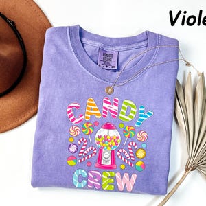 May include: Violet-colored t-shirt with a candy-themed graphic. The design features the words "CANDY CREW" in colorful, bubble letters, surrounded by lollipops, candy canes, and a gumball machine. A gold necklace is draped across the shirt.