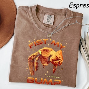 May include: Espresso-colored t-shirt with a crew neck and short sleeves. The shirt features a graphic of a fist bump with the words "FIST MY BUMP" in an orange and red gradient. A silver necklace with a pendant is draped across the shirt.