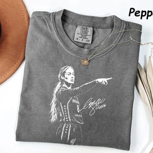 May include: A pepper-colored short-sleeve t-shirt featuring a white graphic print of a basketball player pointing, with a signature and the number 8. The shirt has a Comfort Colors label at the inner neckline, indicating size small, 100% cotton, made in Honduras.