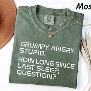 May include: A moss green t-shirt with the text "GRUMPY. ANGRY. STUPID. HOW LONG SINCE LAST SLEEP, QUESTION?" in white. A gold necklace with a pendant is draped across the shirt.