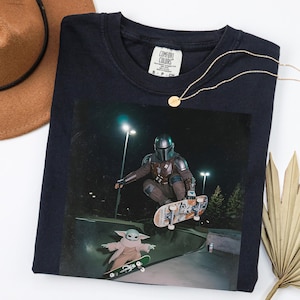 May include: Navy blue long-sleeved t-shirt with a graphic of The Mandalorian and Grogu skateboarding at night. The shirt has a crew neck and a Comfort Colors tag. A gold necklace and a brown hat are also in the image.
