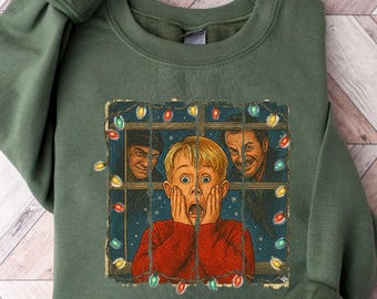 May include: An olive green sweatshirt featuring a graphic from the movie "Home Alone." The graphic shows a young boy with his hands on his face, looking shocked, with two men peering through a window. The image is framed with Christmas lights.