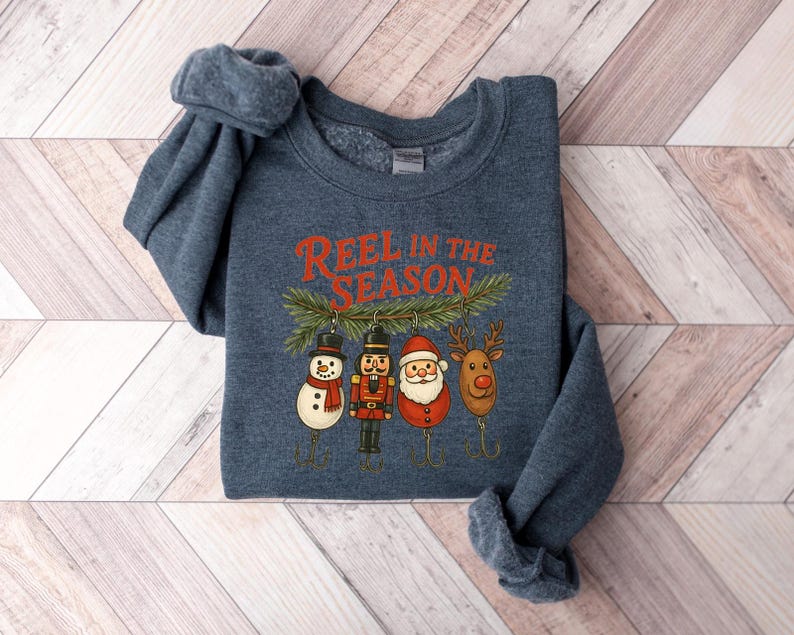 May include: A navy blue sweatshirt with the text "Reel in the Season" in orange. The graphic features a snowman, nutcracker, Santa, and reindeer, each designed as a fishing lure. The sweatshirt is folded on a light wood-grain surface.