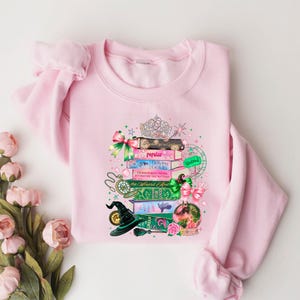May include: A light pink sweatshirt featuring a whimsical design of stacked books, a tiara, and a witch's hat. The books have titles like "Popular" and "The Official Guide". The design includes pink and green bows, a snow globe, and a pocket watch.