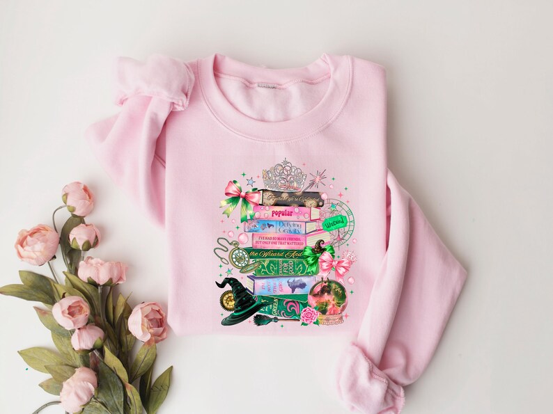 May include: A light pink sweatshirt featuring a whimsical design of stacked books, a tiara, and a witch's hat. The books have titles like "Popular" and "The Official Guide". The design includes pink and green bows, a snow globe, and a pocket watch.