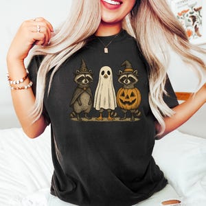May include: Black t-shirt featuring a Halloween-themed graphic. The design includes two raccoons in witch hats and a ghost, all standing together. One raccoon holds a pumpkin. The artwork has a vintage, cartoon style.