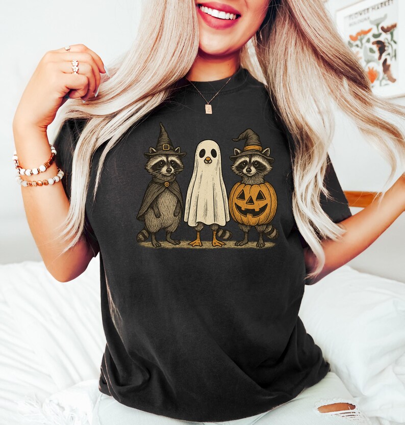 May include: Black t-shirt featuring a Halloween-themed graphic. The design includes two raccoons in witch hats and a ghost, all standing together. One raccoon holds a pumpkin. The artwork has a vintage, cartoon style.