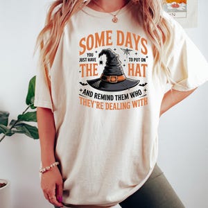 Puede incluir: Camiseta color crema con el texto "SOME DAYS YOU JUST HAVE THE HAT TO PUT ON + AND REMIND THEM WHO THEY'RE DEALING WITH." El dise&ntilde;o incluye un sombrero de bruja, una ara&ntilde;a y murci&eacute;lagos.